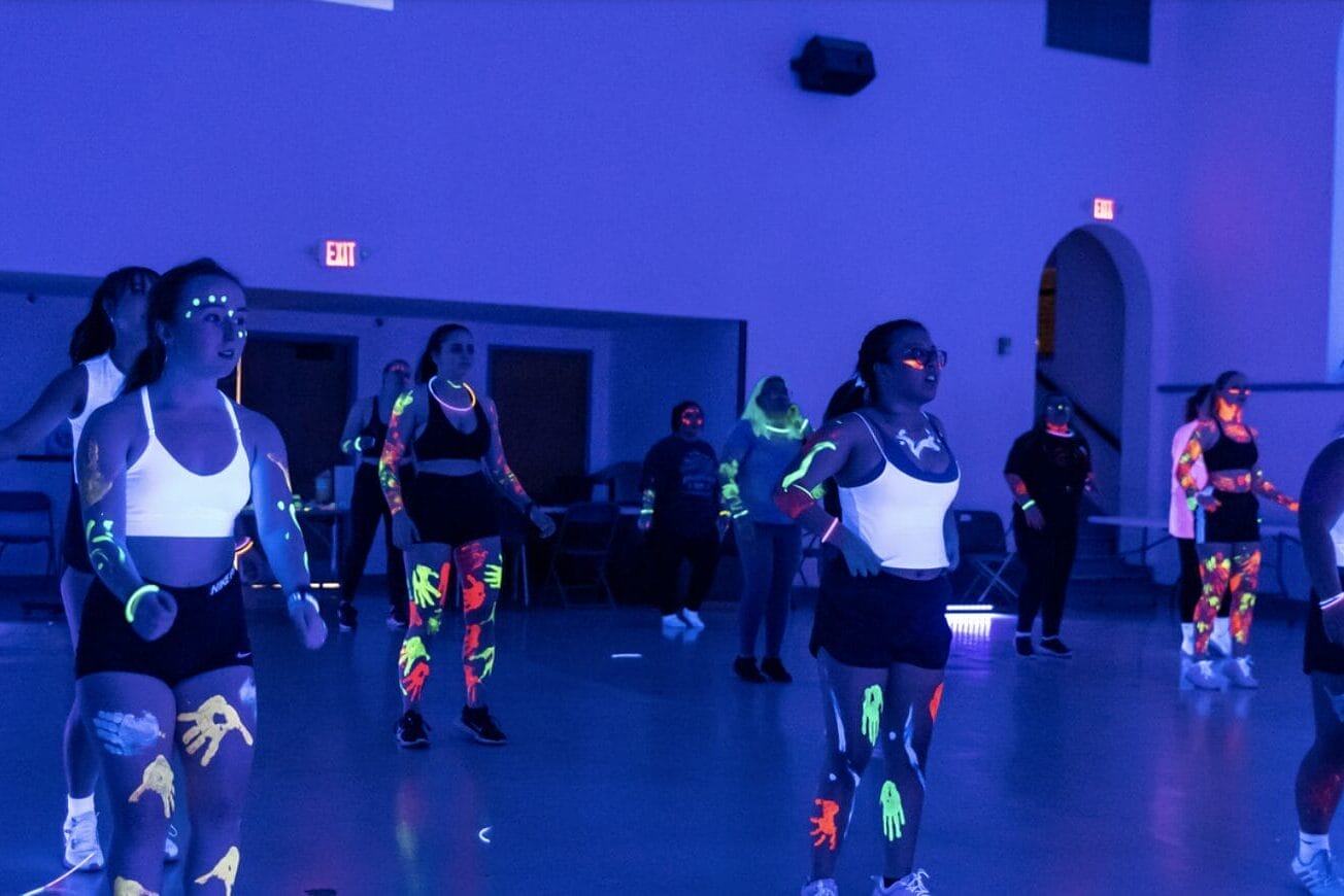People in athletic wear participate in a recreation fitness class under black lights, with neon paint on their faces, arms, and legs glowing brightly. The dimly lit room is filled with a blue and purple sports-inspired hue.